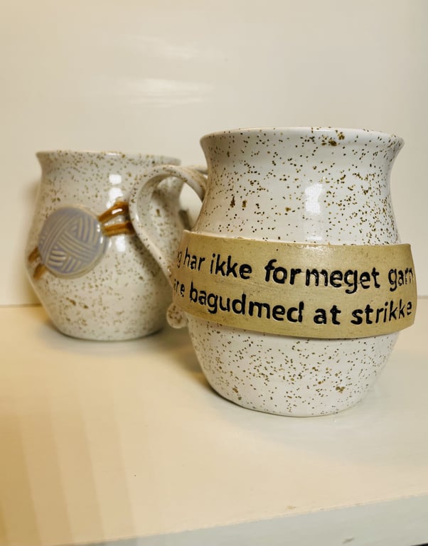 To keramikkrus med beige og brune prikker. Det forreste krus har en beige bånd med dansk tekst og det bagerste krus har et dekorativt skiverelief.