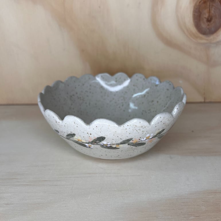 Lille keramisk skål med bølget kant og blomstermotiv på en lys træbakgrund.