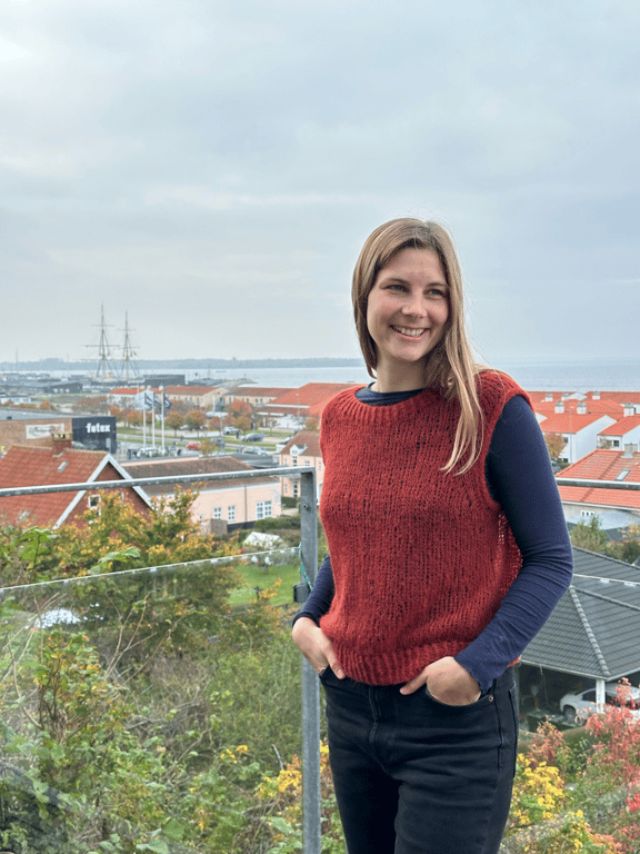 Ung kvinde med langt lyst hår iført rød strikket vest og mørk langærmet trøje står smilende ved gelænder med udsigt til havn, skib og bygninger med røde tage under en grå himmel.