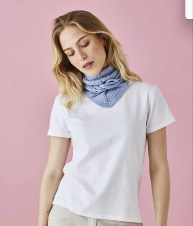 Kvinde med lyst hår iført hvid t-shirt og lyseblåt strikket halstørklæde foran pink baggrund.