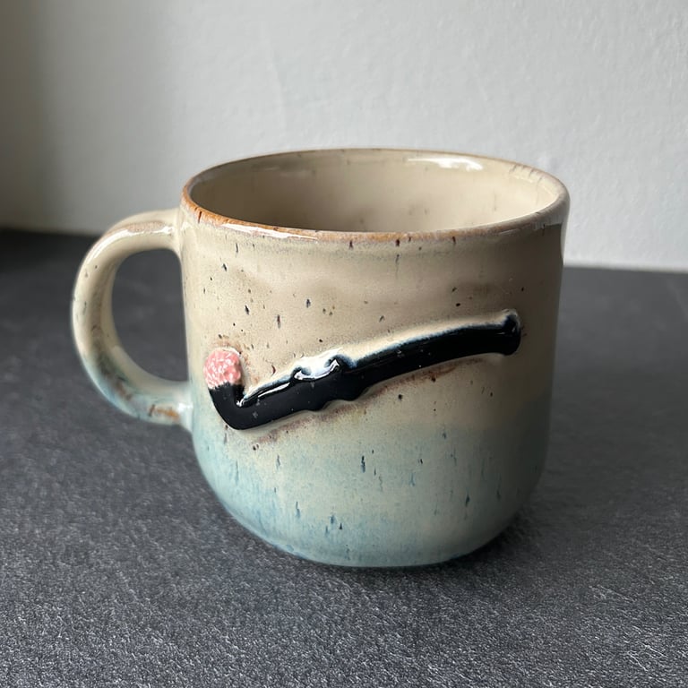 Keramisk krus med håndtag og en pynt i form af en tændt tændstik på ydersiden, placeret på en mørk overflade.