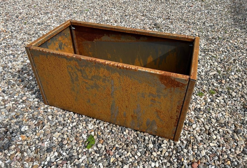 Rustent metalspand eller beholder placeret på en grusbelagt overflade.