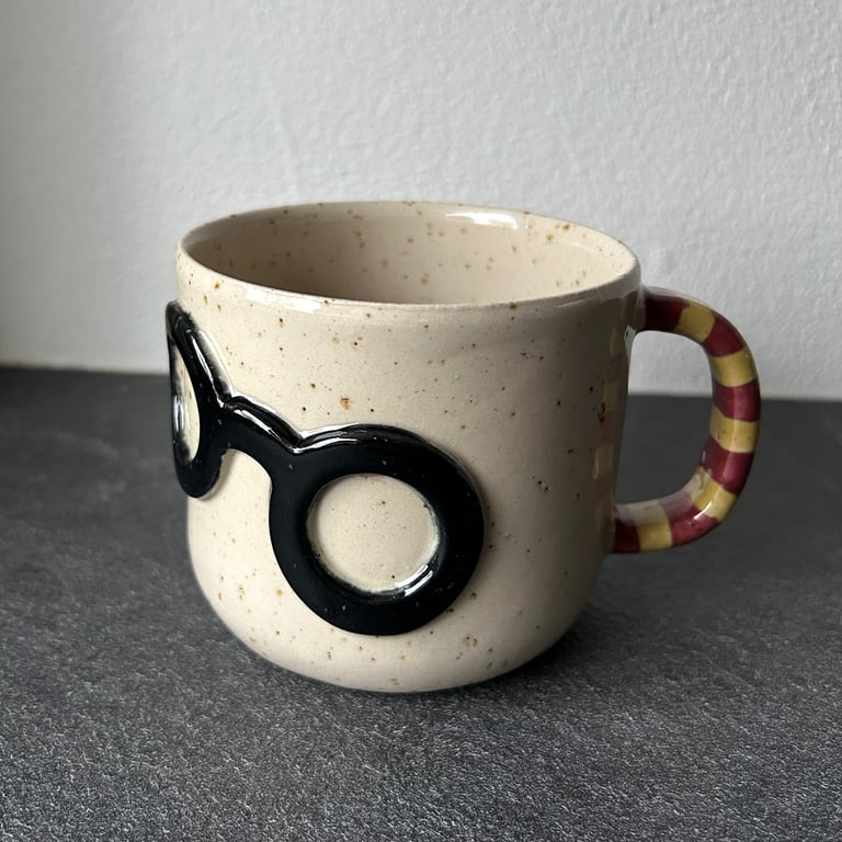 Keramikkrus med sort brillemotiv på forsiden og hank med røde og gule striber.