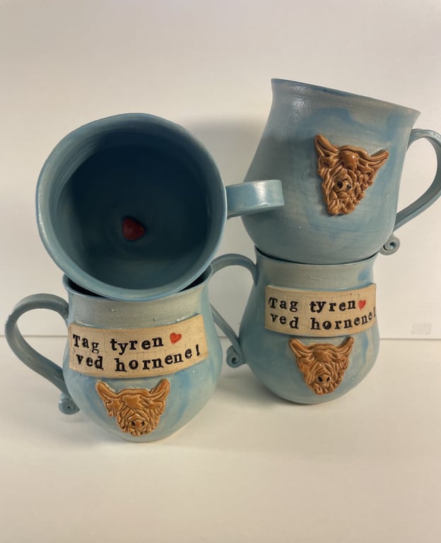 To lyseblå keramikkrus med brune hornet tyrehoveder og teksten "Tag tyren ved hornene" på forsiden. Et krus står på hovedet med et lille rødt hjerte i bunden.