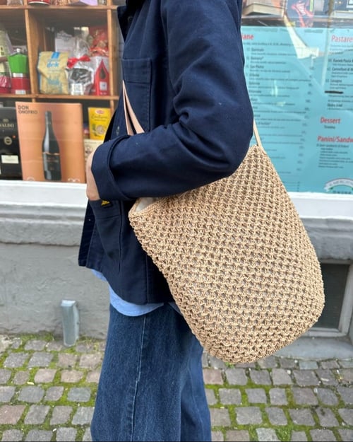 Person med mørk blå jakke og blå jeans bærer en strikket, beige taske over skulderen foran en butik med glasvindue. (1)
