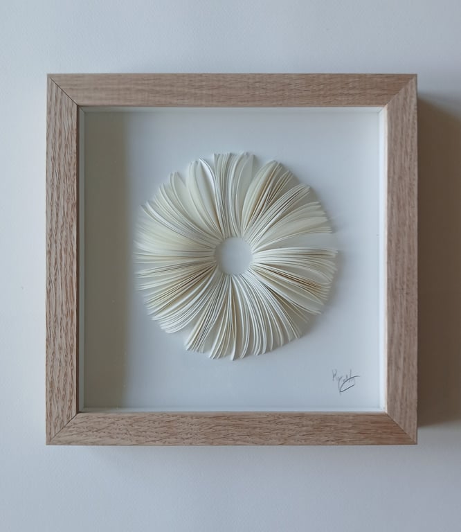 Rammet vægkunst med foldede sider af en bog arrangeret i en cirkel med et hul i midten, på hvid baggrund.