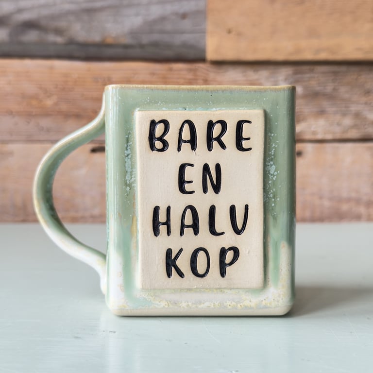 Grøn keramikkop med teksten "Bare en halv kop" på fronten, stående på en lys bordplade med træbaggrund.