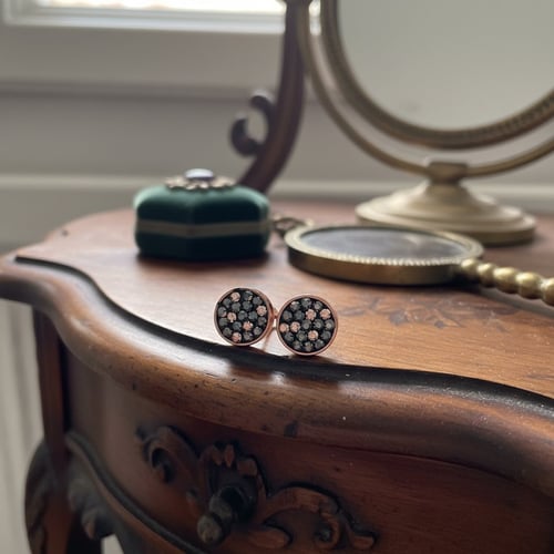Et par runde ørestikker med nitter i sort og rosa metal placeret på kanten af et træmøbel, med en vintage spejl og et smykkeskrin i baggrunden. (1)