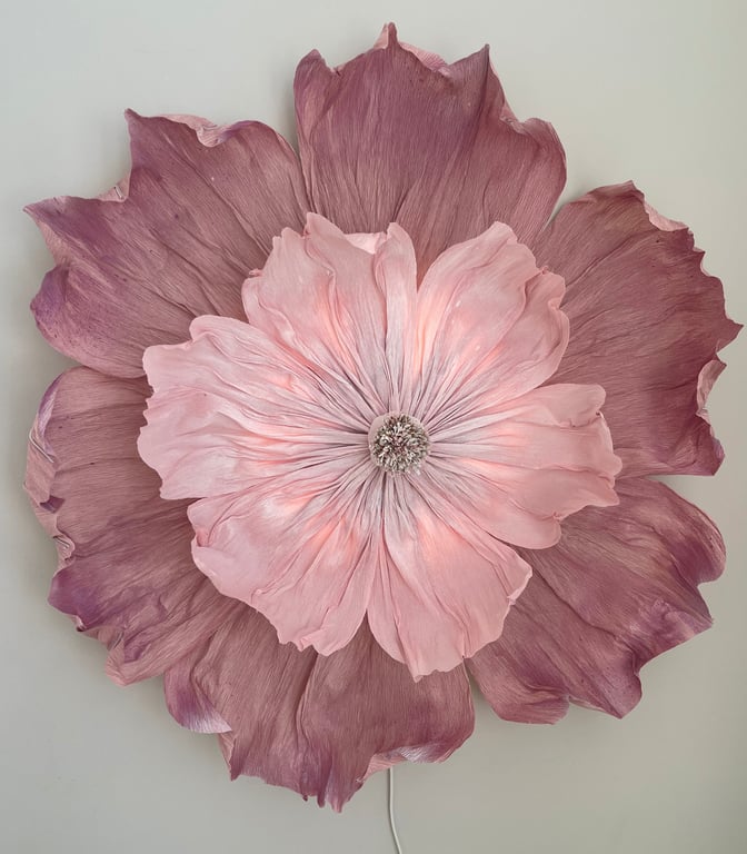 Stor vægdekoration formet som en blomst med lyserøde og lilla kronblade mod en lys væg.