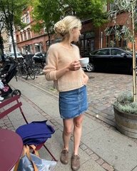 En kvinde med lyst hår i en lav knold, iført beige striktrøje og jeansnederdel, står på en fortov i en by og holder en hvid kop. Bag hende ses cykler, biler og træer langs gaden. (3)