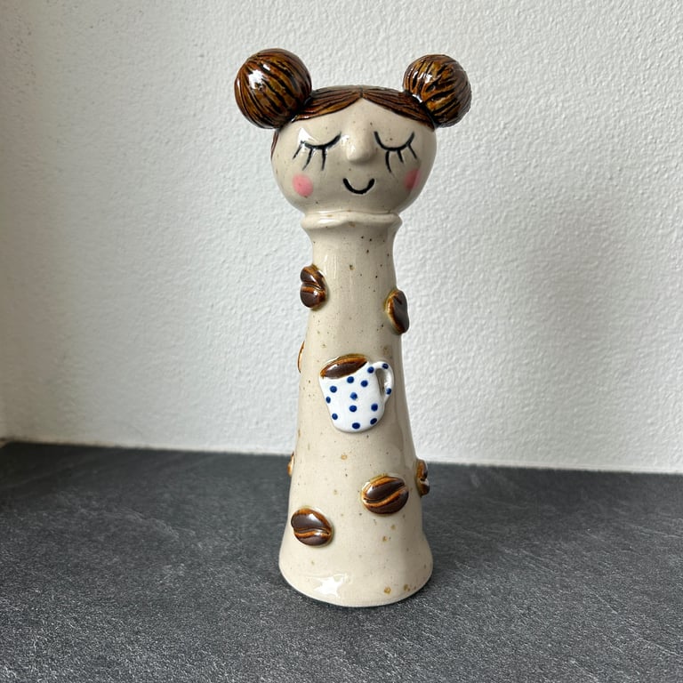 Keramisk figur med lukkede øjne, rødroser kinder, brunt hår i to knolde, dekoreret med kaffebønner og en hvid kaffekop med blå prikker, stående på en mørk bordplade mod en hvid væg.