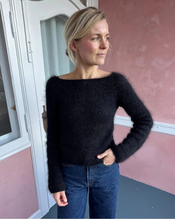 Kvinde iført sort, langærmet, strikket sweater og blå jeans, stående ved en lyserød væg og en hvid dør.