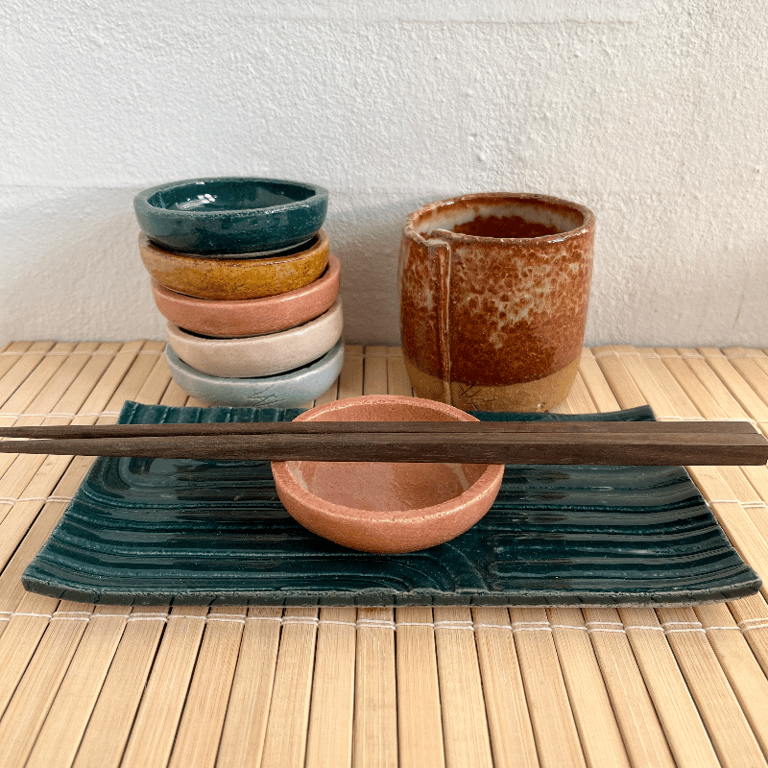 Keramisk japansk spisebord med spisepinde, små skåle i forskellige farver og en rustik kop på bambusmåtte.