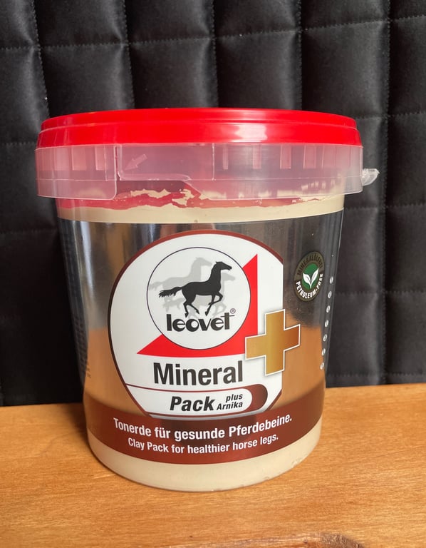 Plastbeholder med rød låg indeholdende Leovet Mineral+ Pack lerpakning til sunde hesteben, stående på et bord foran en sort polstret baggrund.