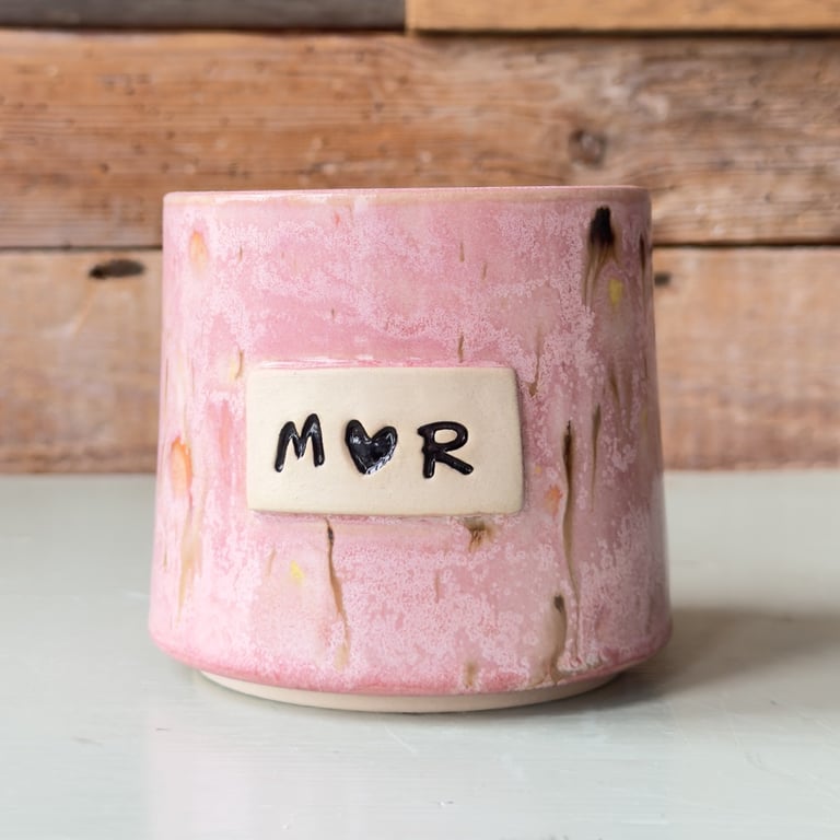 Keramikkop med lyserød glasur og etiketten "M♥R" på forsiden, placeret på en lys overflade med træbaggrund.