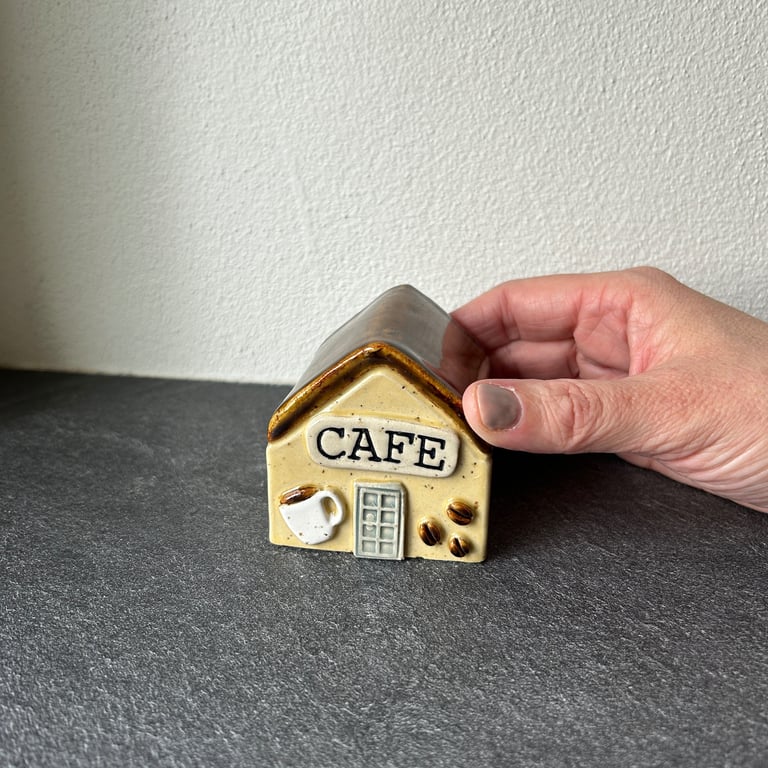 Hånd der holder en lille keramikfigur formet som et caféhus med teksten "CAFE", en kaffekop og kaffebønner foran.