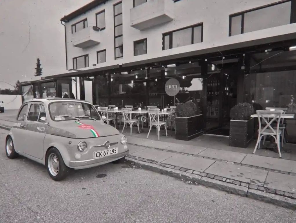Sort-hvid billede af en klassisk Fiat 500 parkeret foran en restaurant med udendørs caféområde og hvidmalede stole.