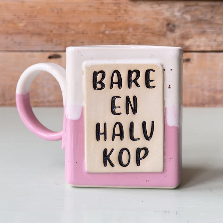 Kop med tekst "Bare en halv kop" i sort skrift på en lyserød og hvid baggrund, stående på en hvid overflade med en træbaggrund.