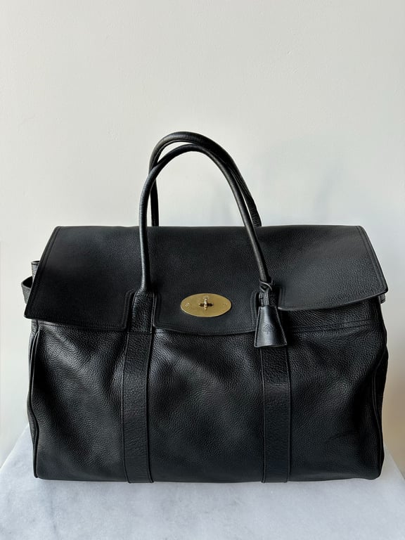 Sort lædertaske med to håndtag og guldfarvet drejelås foran, placeret på hvid overflade foran hvid væg.