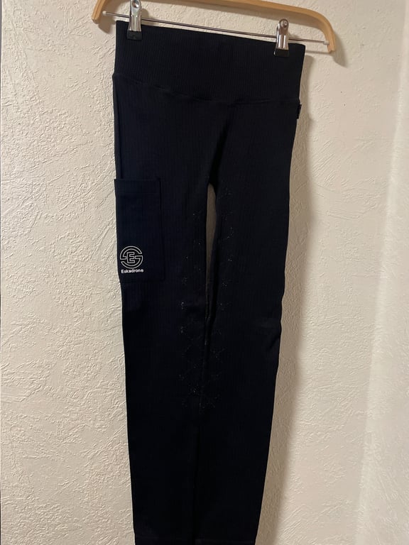 Sort leggings med høj talje og lomme på siden, hængende på en bøjle mod en væg.