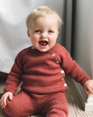 En smilende baby iført en strikket, rustrød trøje og bukser, siddende indendørs med et lyst gardin i baggrunden. (2)
