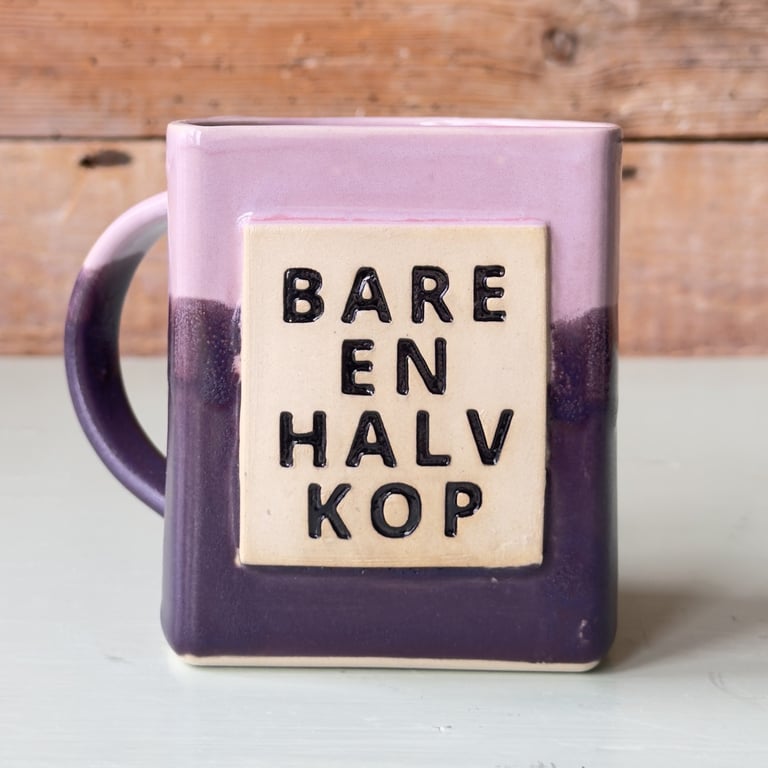 Lilla og lyserød firkantet keramikkrus med teksten "BARE EN HALV KOP" på en beige firkant på fronten, stående på en lys overflade med en træbaggrund.
