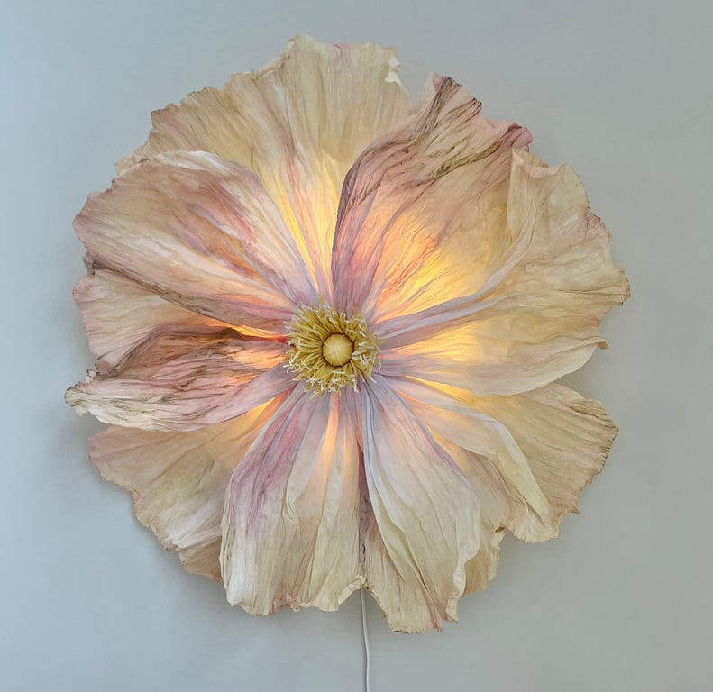 Stor blomst formet væglampe med sarte, bølgede kronblade i lyse nuancer af creme, rosa og gul, oplyst indefra mod en lys væg.