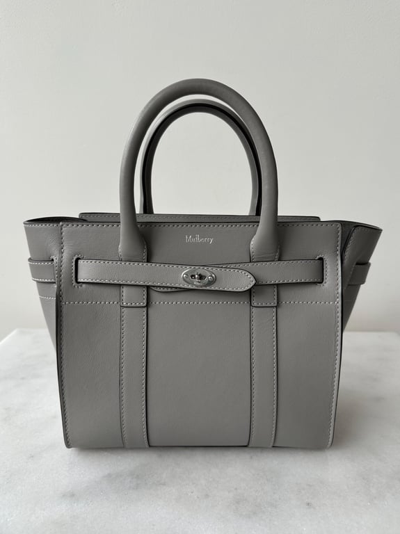 Grå lædertaske med to håndtag og metalspænde på forsiden, placeret på hvid overflade mod en lys baggrund.