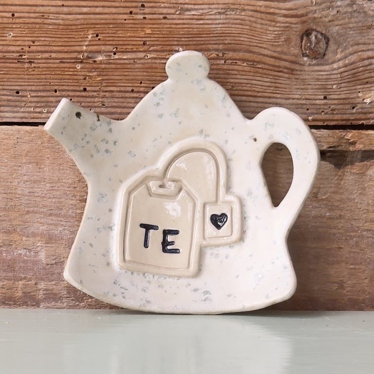 Keramisk tekandeformet underkop med en udstikket tebrevsfigur og teksten "TE", stående foran en rustik trævæg.