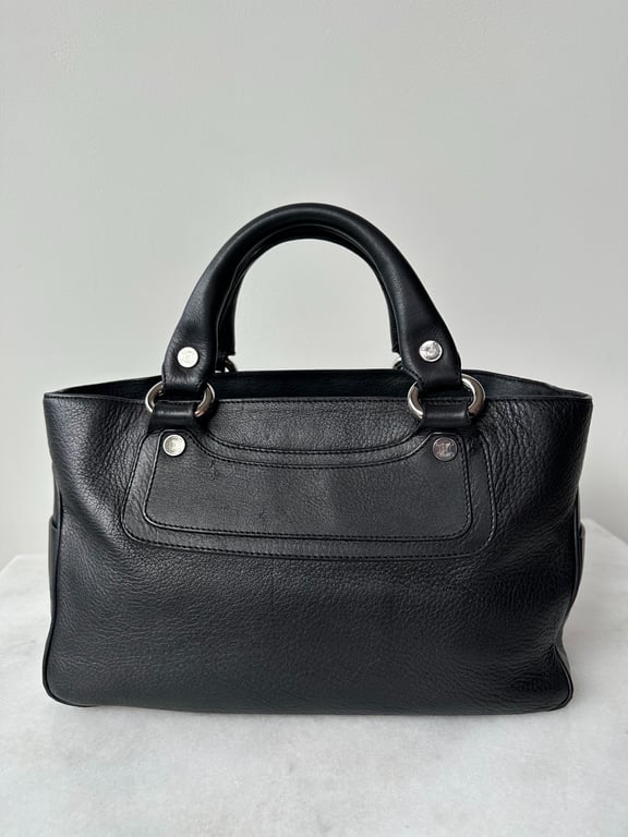 Sort lædertaske med korte håndtag og sølvfarvede spænder, placeret på en hvid overflade mod en grå baggrund.