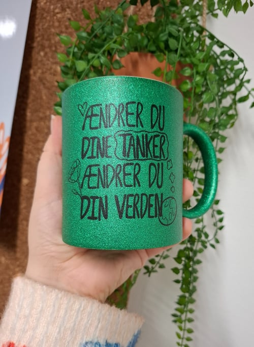 Hånd, der holder en glitrende grøn krus med sort tekst på dansk, der siger "Ændrer du dine tanker, ændrer du din verden", med tegninger af et hjerte, en rose og en jordklode, foran en grøn plante. (1)