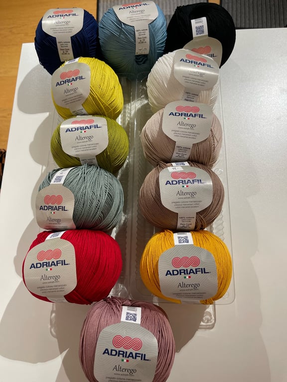 Billede af 13 nøgler garn i forskellige farver fra mærket Adriafil Alterego, arrangeret i to kolonner på en hvid overflade.