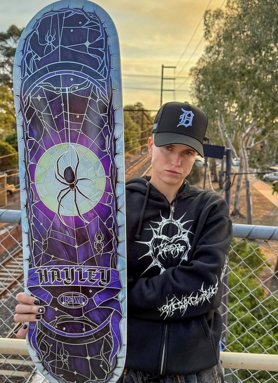 Ung person iført sort hættetrøje og kasket, der holder et skateboard med mørkelilla og sort spindelvæv-design med en edderkop i midten.