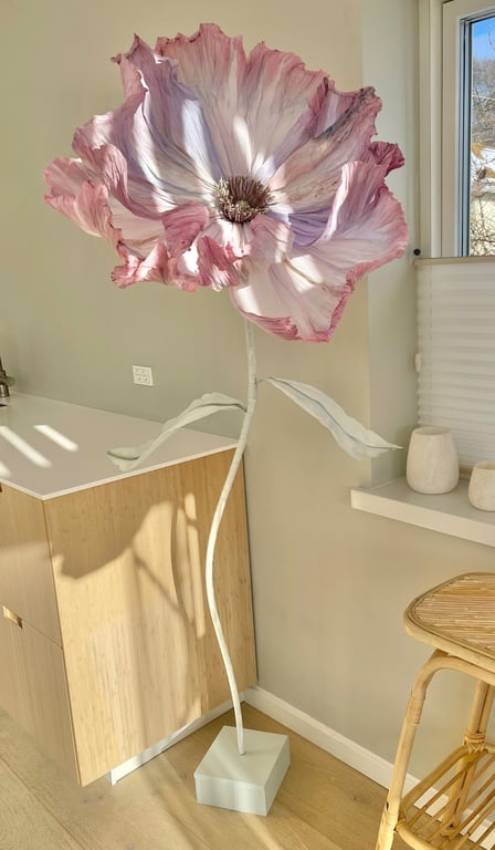 Stor kunstig blomst i lyserøde og hvide nuancer, placeret i et lyst rum med træmøbler og et vindue i baggrunden.