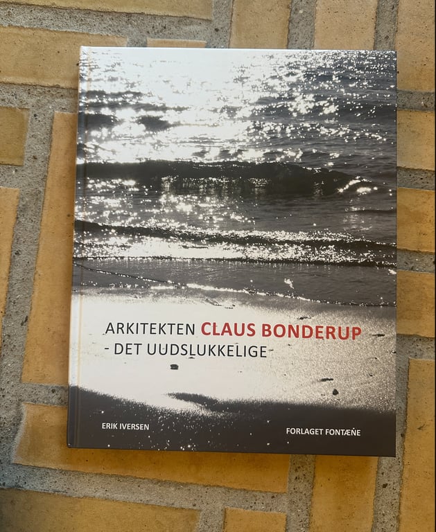 Bog med titlen "Arkitekten Claus Bonderup - Det uudslukkelige" af Erik Iversen, liggende på gulv med gulbrune fliser. Forsiden viser et sort-hvidt billede af vand med sollys, der reflekterer.