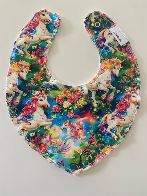 Bibs med farverigt unicorn-print og blomster på blå og grøn baggrund.