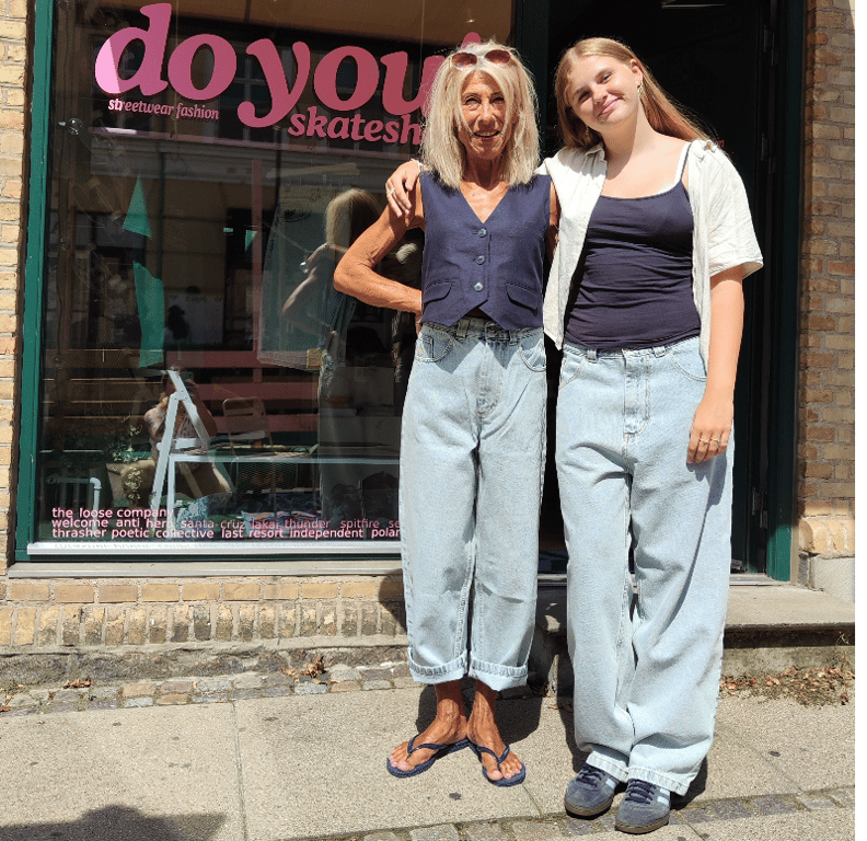 To kvinder, en ældre og en yngre, står tæt sammen foran en butik med skater-tøj, begge iført lyse baggy jeans og mørke toppe, smilende i sollys.