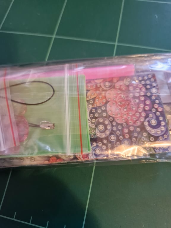 En plastikpose med mindre lynlåsemballager indeholdende perler og et smykkewire med en lås, lagt på en grøn skæreplade.