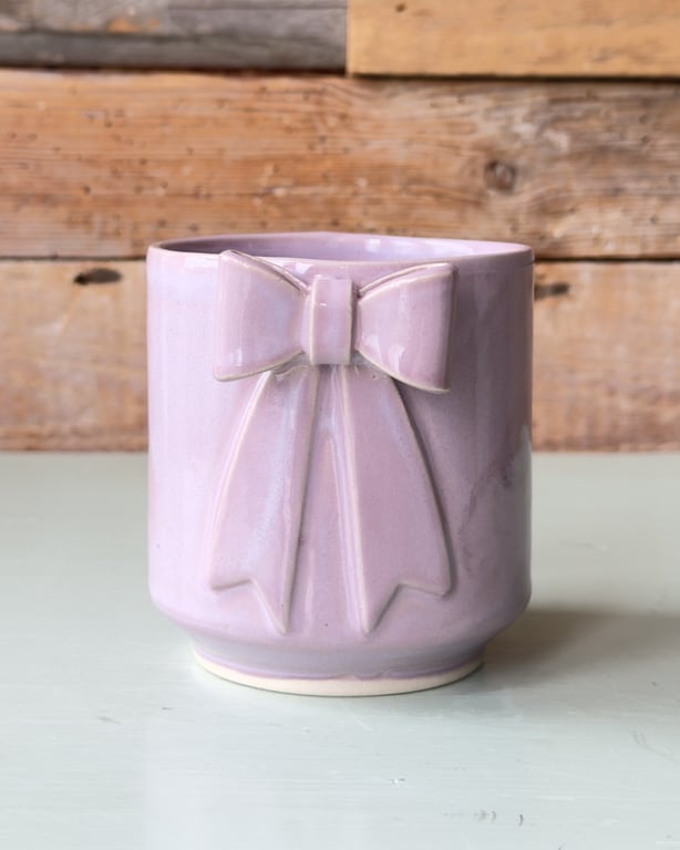 Lilla keramik krus med dekorativ sløjfe på fronten, placeret på lys overflade med træbaggrund.