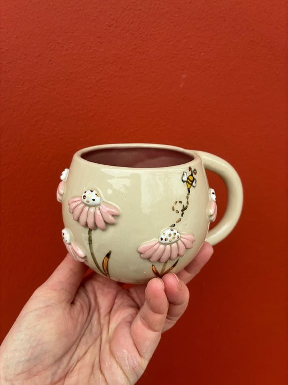 En hånd holder en beige keramikkrus med 3D-udsmykning af lyserøde blomster og en lille bi mod en rød baggrund.
