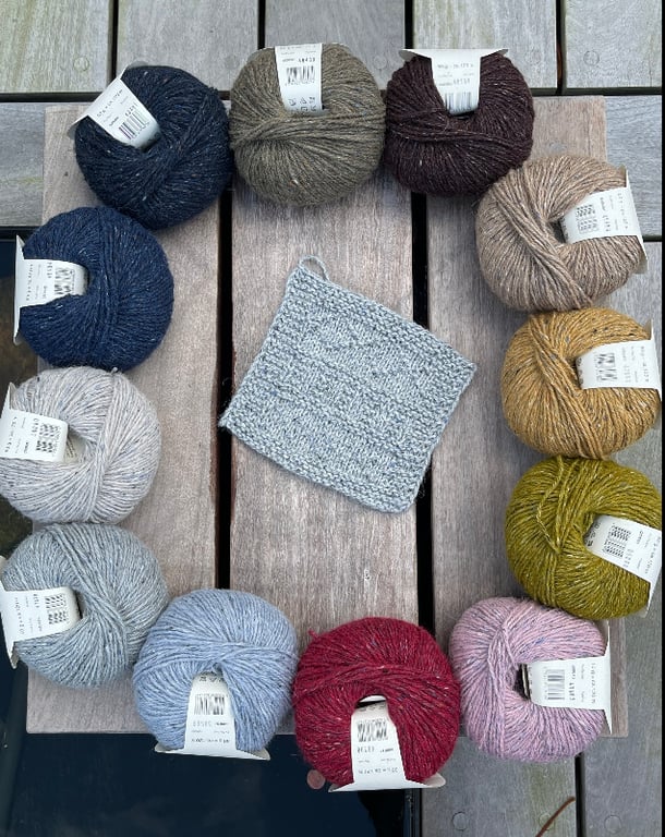 Billede af flere garnnøgler i forskellige farver arrangeret i en cirkel på et træbord med et lille strikket firkant i midten.