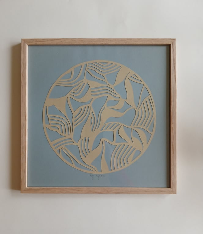 Abstrakt cirkulært kunstværk med beige og blå mønstre i en træramme, hængende på en hvid væg.