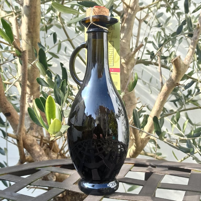 Sort glasflaske med håndtag og korkprop, placeret på metalbord foran oliventræ.