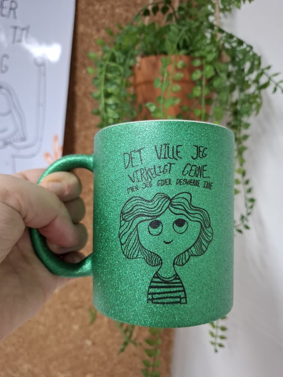Grøn glitrende kop med tegning af et smilende ansigt og dansk tekst, holdt i en hånd, med en plante og opslagstavle i baggrunden.