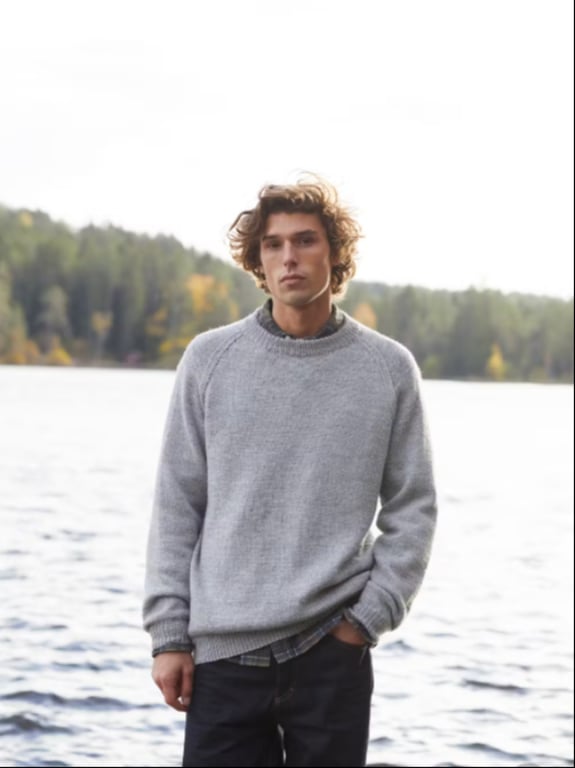 Ung mand iført grå sweater står ved en sø med skov i baggrunden. (3)