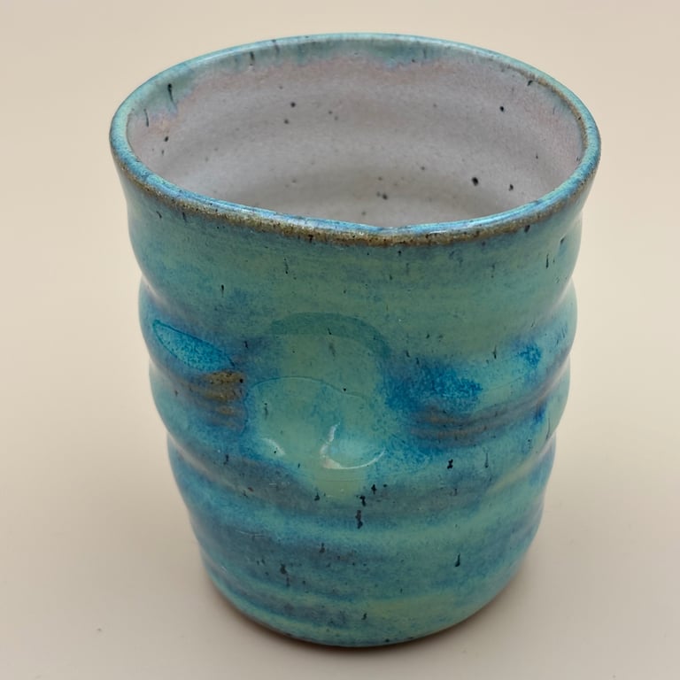 Blågrøn keramikkrus med bølget struktur og glaseret overflade, stående på beige baggrund.