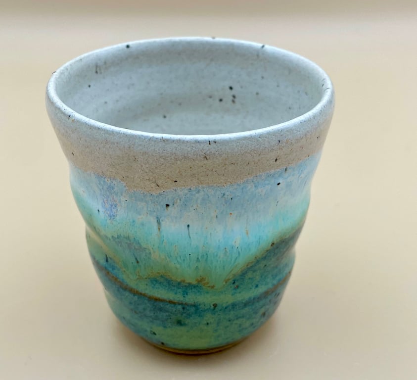 Keramikkrus med bølget form og glasur i blå, grønne og grå nuancer på beige baggrund.