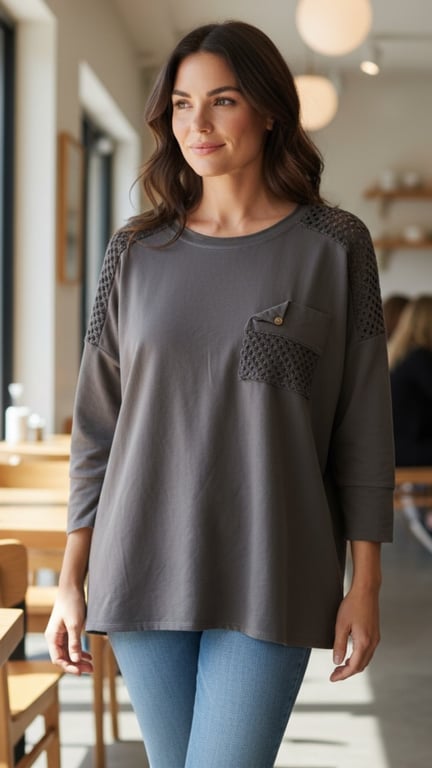 Ung kvinde iført grå bluse med mønstret lommedetalje og lyseblå jeans, står i lyst caféindretning.