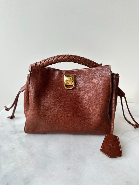 Brun lædertaske med flettet håndtag og guldfarvet spænde, placeret på marmorbaggrund.