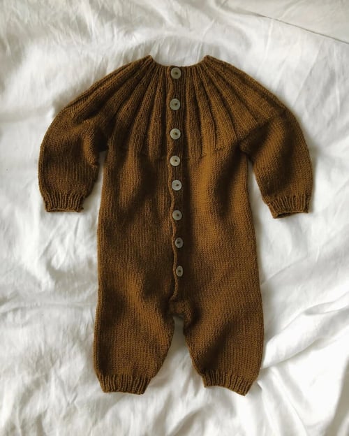 Brun, strikket babyheldragt med knapper ned foran, lagt på hvidt stof. (1)